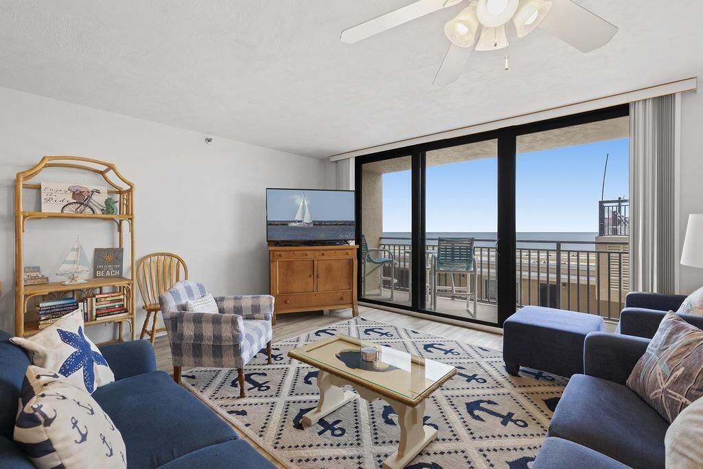 A living room with a large window overlooking the sea, a television, and a bookshelf.