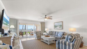 A spacious living room with striped sofas, a coffee table, and a ceiling fan. There is a large window offering a view of the ocean.