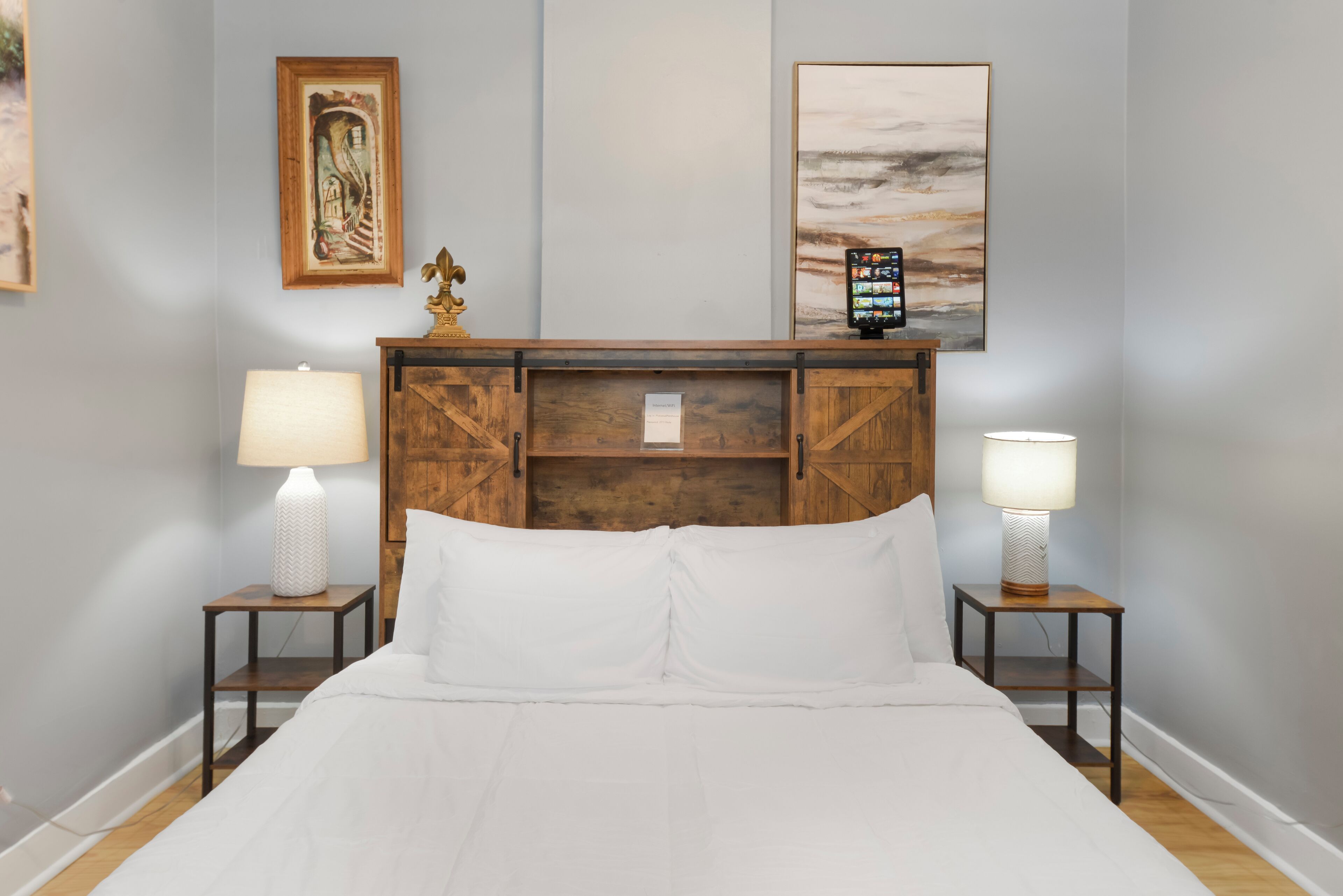 Uma cama bem arrumada com lençóis brancos, duas mesas de cabeceira com abajures, um painel de cama de madeira e duas obras de arte emolduradas na parede.