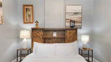 Uma cama bem arrumada com lençóis brancos, duas mesas de cabeceira com abajures, um painel de cama de madeira e duas obras de arte emolduradas na parede.