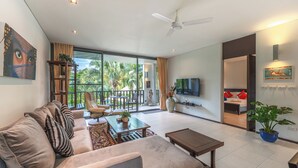 A modern living room with a sofa, coffee table, and TV. There is a balcony with a view of palm trees. - C215 Bang Tao Beach Garden F21 (Choeng Thale)