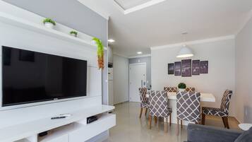 Una sala de estar moderna con un televisor de pantalla plana, un sofá, un comedor con sillas estampadas y una cocina al fondo.