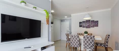 A modern living room with a flat-screen TV, a sofa, a dining area with patterned chairs, and a kitchen in the background.