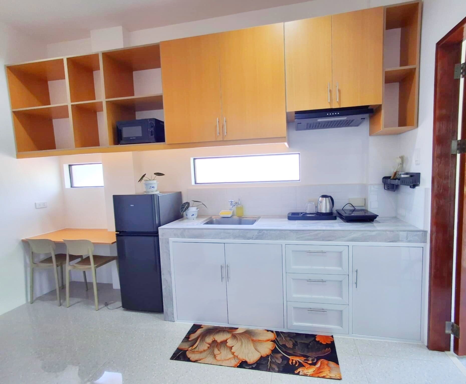 A compact kitchen with white cabinets, a black refrigerator, a sink, and a countertop with various items. There is a window above the sink and a door to the right.