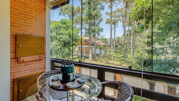 A balcony with a glass table, chairs, and a view of trees and buildings. - Charming Apt in the Heart of Gramado! Rln0105 (Gramado)