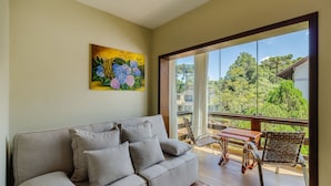 A living room with a grey sofa, a painting of flowers, and a balcony with a wicker chair and table. - Charming Apt in the Heart of Gramado! Rln0105 (Gramado)