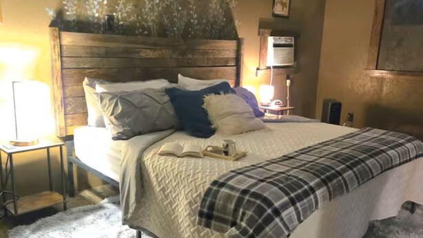 A bedroom with a bed, a ceiling fan, a nightstand, a lamp, and a wall-mounted air conditioner. - The Hideaway - Private Mini-Farm Retreat for two (Evensville)