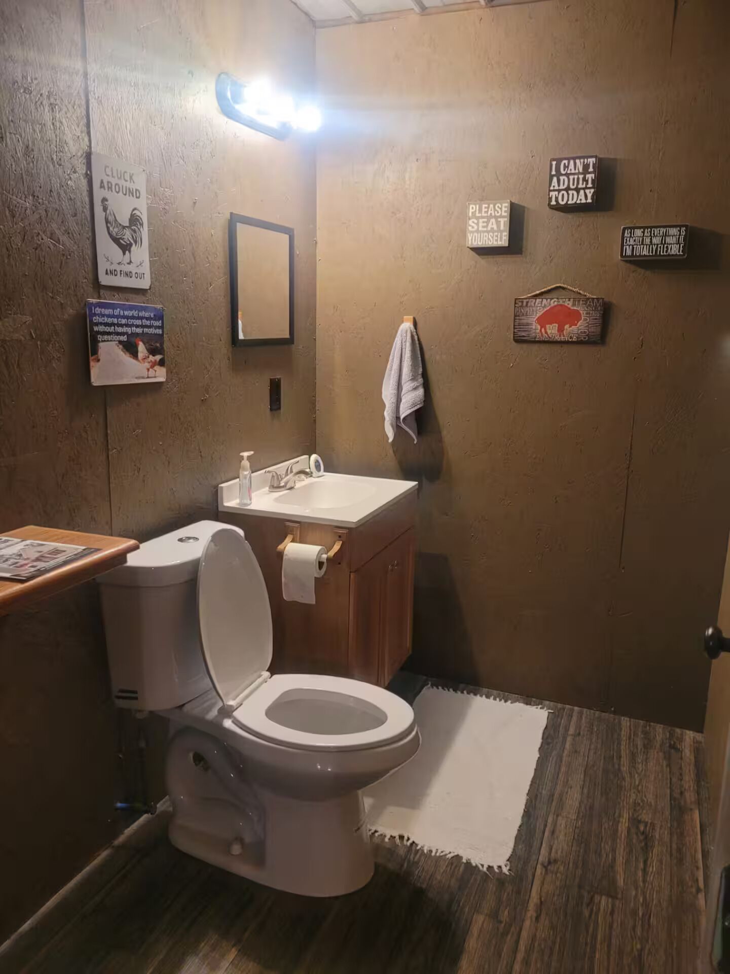 Un baño pequeño con inodoro, lavabo y espejo. Hay varios carteles en las paredes, incluyendo uno que dice 'I can't adult today'.
