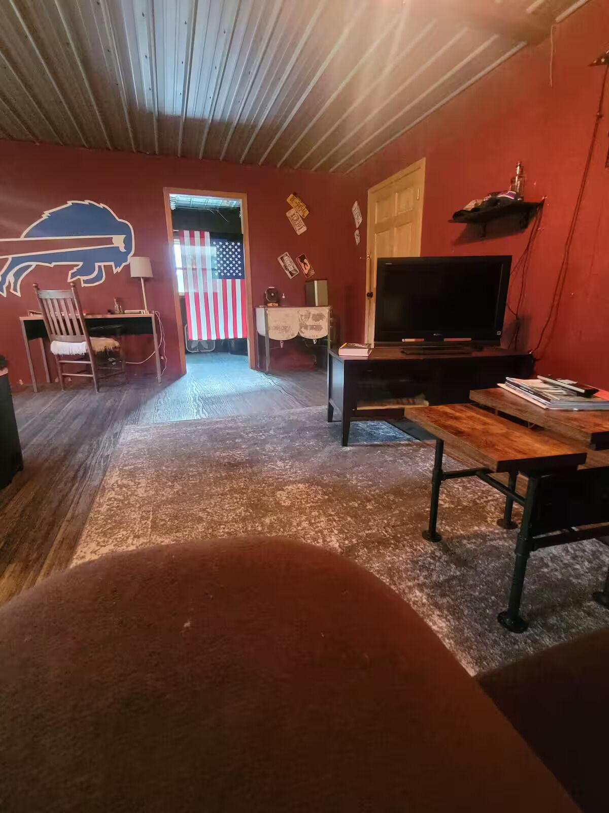 A room with a wooden ceiling, a TV, a desk, a chair, a wooden table, and an American flag.