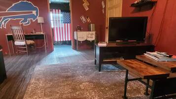 A room with a wooden ceiling, a TV, a desk, a chair, a wooden table, and an American flag.