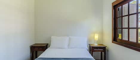 Una cama bien hecha con sábanas blancas, una mesita de noche con lámpara y una ventana con contraventanas.
