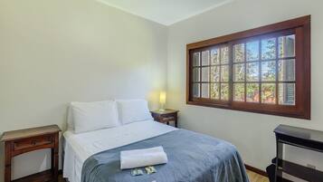 Quarto com cama, mesa de cabeceira, luminária, janela com persianas e escrivaninha.