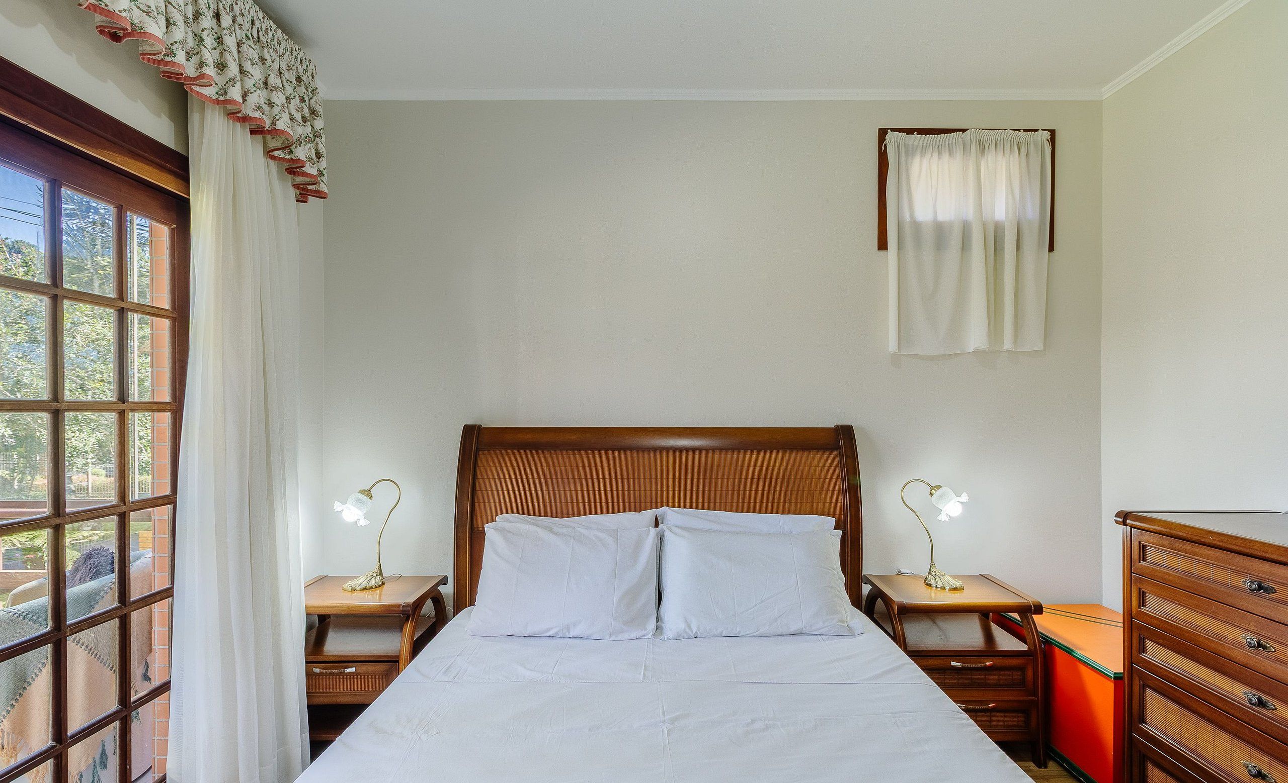 Ein Zimmer mit einem Holzbett, zwei Nachttischen mit Lampen, einem Fenster mit Vorhängen und einer kleinen Holzkommode.