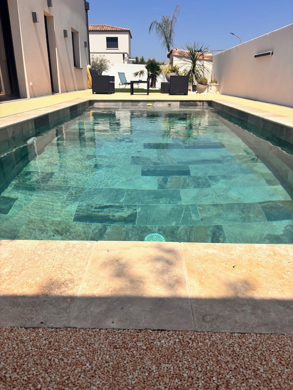 A clear swimming pool with tiled edges, surrounded by a pebble border, and adjacent to a building with a white facade and dark windows.