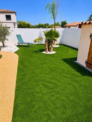 A well-maintained artificial lawn with palm trees and a lounge chair, surrounded by a white wall and a building with a tiled roof.
