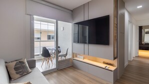 A modern living room with a flat-screen TV mounted on the wall, a sofa with a leaf-patterned pillow, and a sliding glass door leading to an outdoor area with a table and chairs. - Modern apt With Pool and hot Tub! Rhl0305 (Florianópolis)