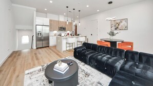 Una cocina moderna con una isla central, un comedor con sillas color naranja y un sofá de cuero negro en una amplia sala de estar.