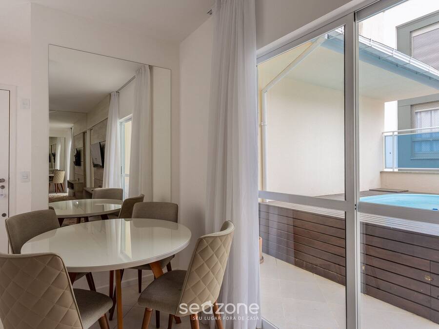 Uma sala de jantar moderna com mesa de vidro e cadeiras acolchoadas, um espelho grande e vista para uma piscina e área externa.