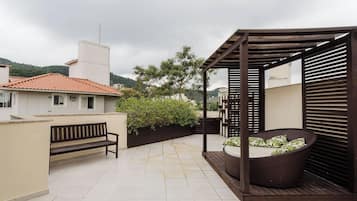 A rooftop patio with a wooden pergola, a wicker chair with cushions, a bench, and a tiled floor.