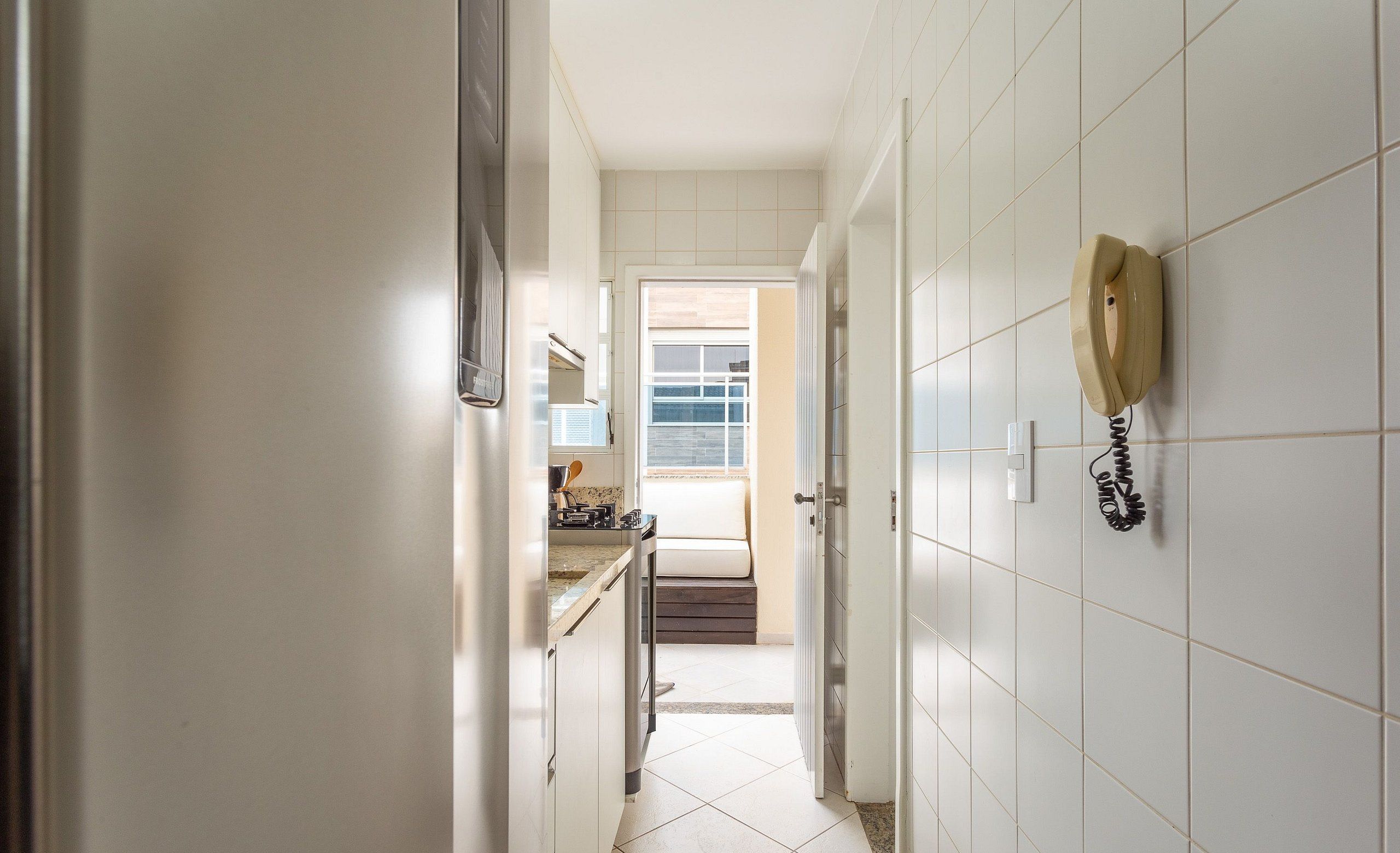 A narrow hallway with white tiled walls, a kitchen area with a stove and countertop visible, and a telephone mounted on the wall.