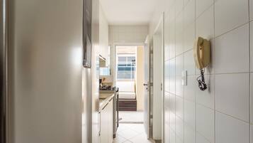 A narrow hallway with white tiled walls, a kitchen area with a stove and countertop visible, and a telephone mounted on the wall.