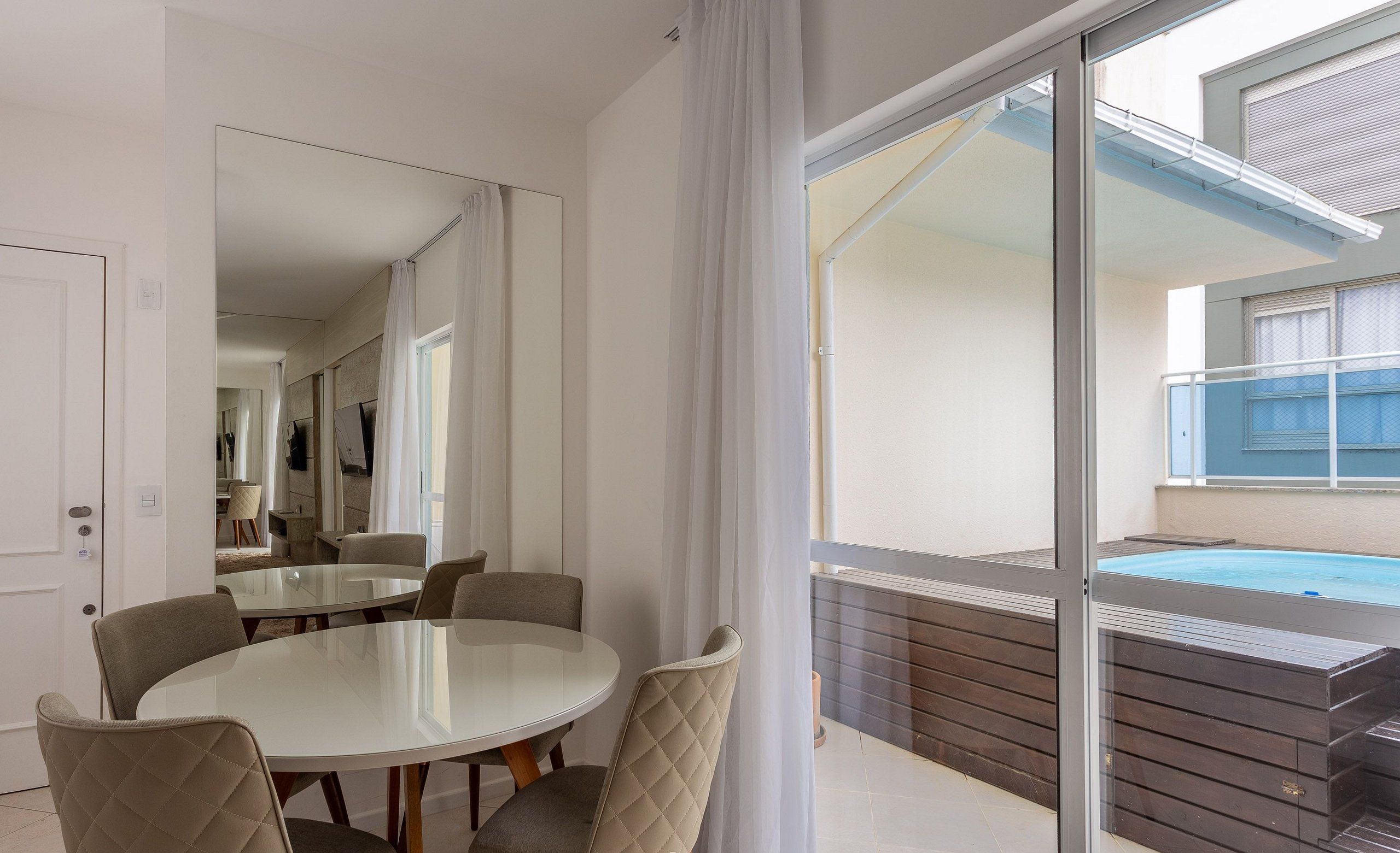 Un comedor moderno con mesa de vidrio y sillas acolchadas, un espejo grande y vistas a una piscina y zona exterior.