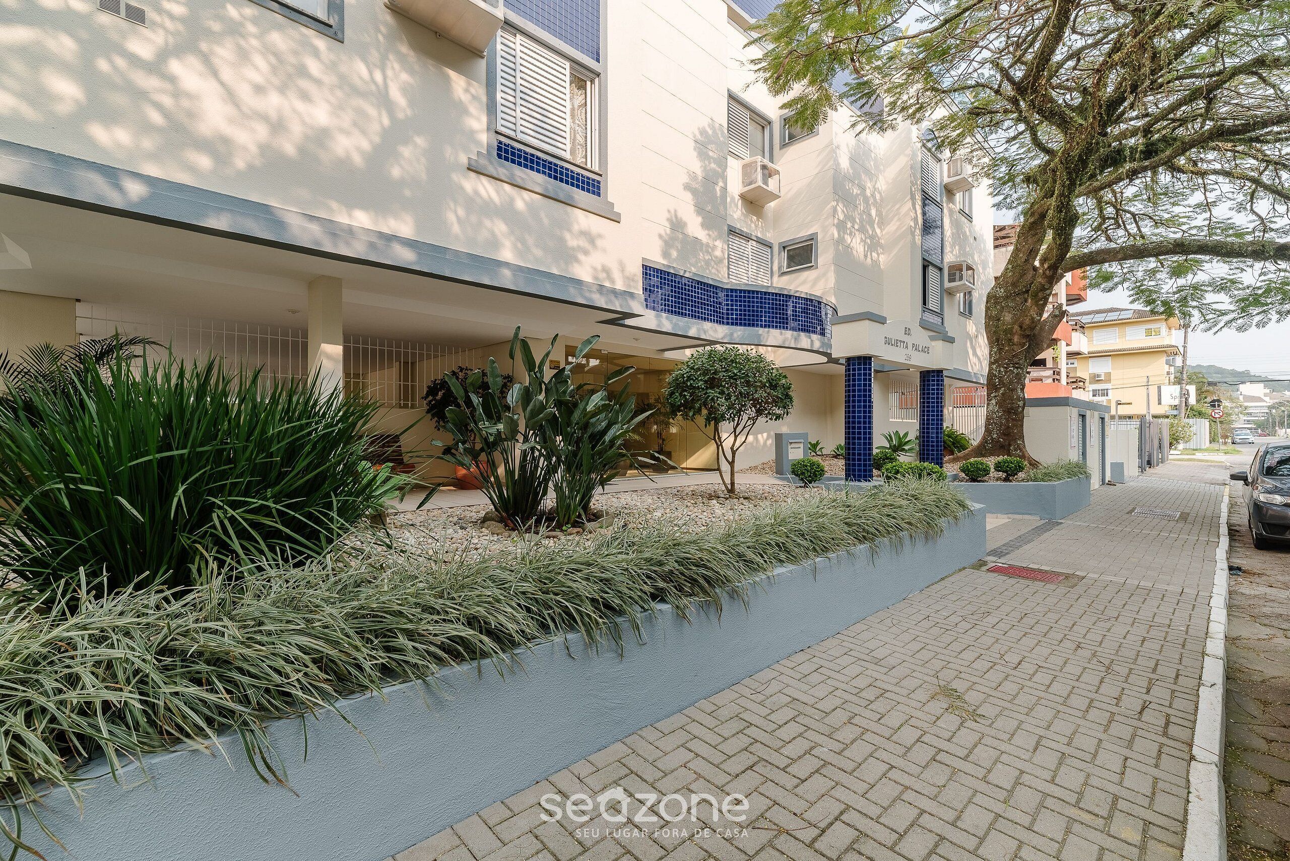 A residential building with a modern design, featuring a balcony with blue tiles, a small garden area with various plants, and a paved walkway.