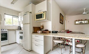 A kitchen with white cabinets, a microwave, a refrigerator, a stove, and a sink. There is a dining area with wicker chairs and a table. - 2 b ap, 400m the Beach, Jurere Downtown Rgp208 (Florianópolis)