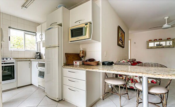 A kitchen with white cabinets, a microwave, a refrigerator, a stove, and a sink. There is a dining area with wicker chairs and a table.