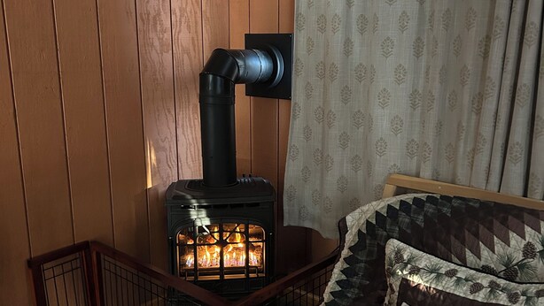 A cozy room with wooden walls, a wood-burning stove, a bed with patterned bedding, and a window with a curtain.