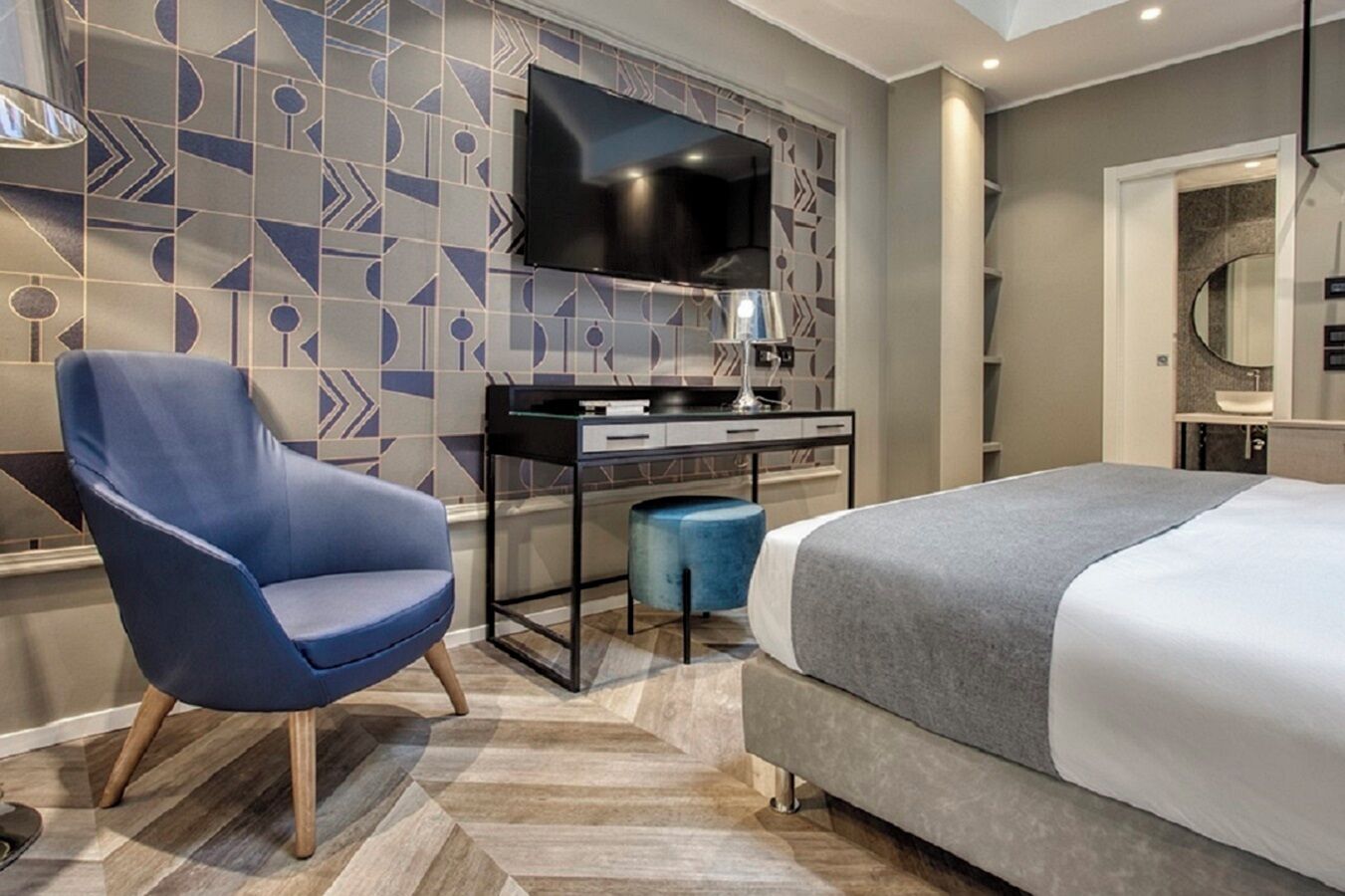 A modern hotel room with a large bed, a desk with a flat-screen TV, a blue armchair, and a patterned wall.