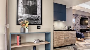 Un soggiorno moderno con pavimento in parquet a spina di pesce, un tavolino consolle blu con libri e candele, un quadro di grandi dimensioni e un angolo cottura con mobili scuri e un televisore.