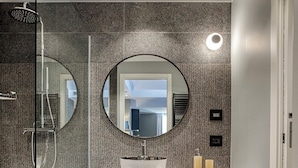 Un bagno moderno con box doccia in vetro, uno specchio rotondo sopra il lavabo e un pavimento in legno a spina di pesce.