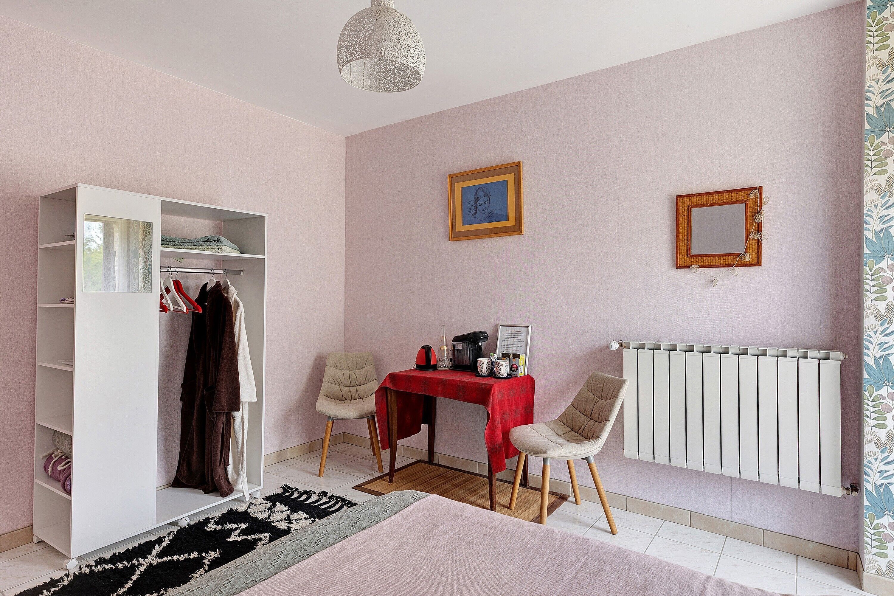 Une chambre à coucher avec une armoire, deux chaises, une table recouverte d’une nappe rouge, un radiateur et un luminaire suspendu.