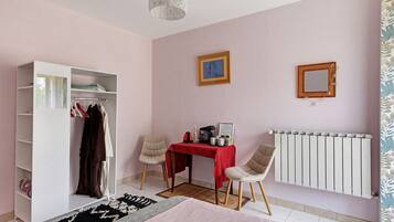 Une chambre à coucher avec une armoire, deux chaises, une table recouverte d’une nappe rouge, un radiateur et un luminaire suspendu.