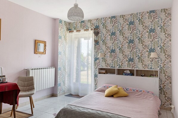 Un dormitorio con una cama, una silla, una mesa, un radiador, una ventana con cortinas y una pared con estampado floral.