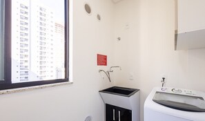 A laundry room with a washing machine, a sink, and a window overlooking a high-rise building. - Apart With Barbecue 600m From the Beach Rgoa701 (Itapema)