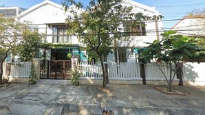 Un edificio de dos plantas con fachada blanca, entrada protegida por una reja y un pequeño jardín con árboles y arbustos.