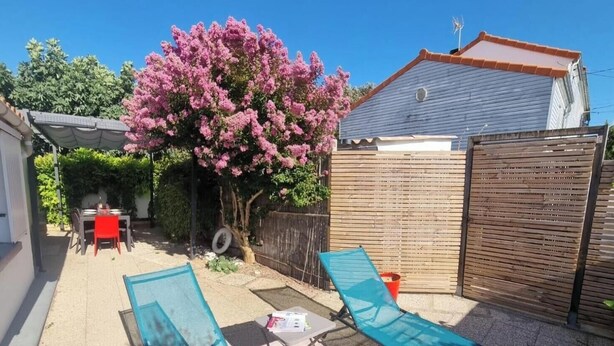 Un jardin avec une table et des chaises de patio, un fauteuil bleu, un arbre aux fleurs roses et une clôture en bois.