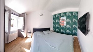 Une chambre à coucher comprenant un lit, une fenêtre, un radiateur, une table de chevet et un téléviseur fixé au mur.
