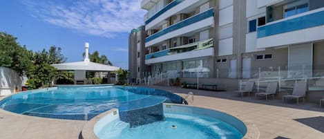 A swimming pool area with a circular hot tub, a covered seating area, and a multi-story building with balconies.