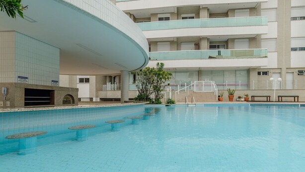En swimmingpool med en buet bygning i baggrunden, flere balkoner og krukker med planter.