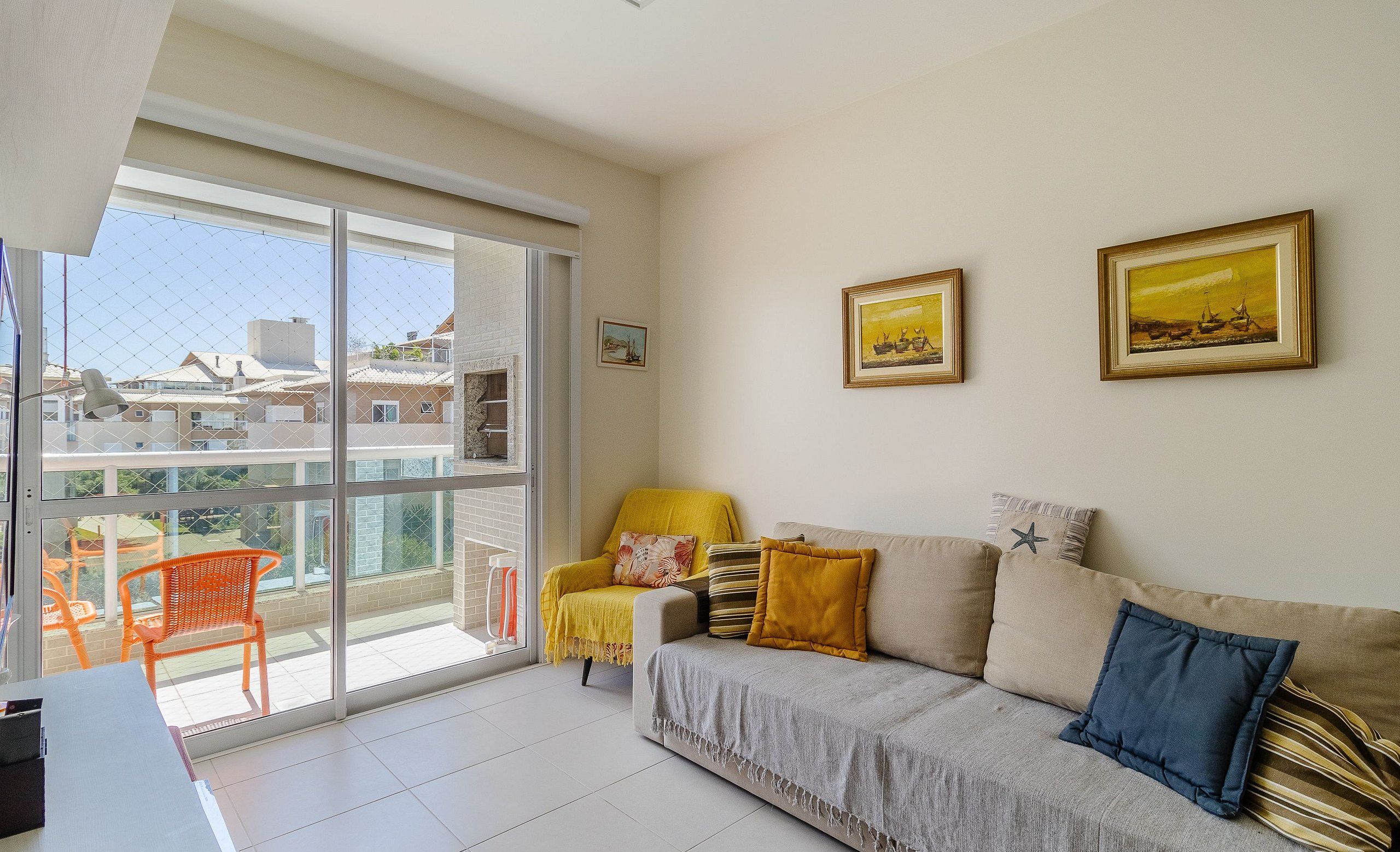 A living room with a sofa, armchairs, and a dining table. There is a large window with a view of buildings and a balcony.