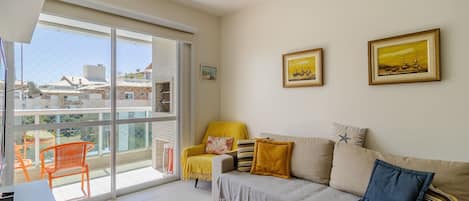 A living room with a sofa, armchairs, and a dining table. There is a large window with a view of buildings and a balcony.