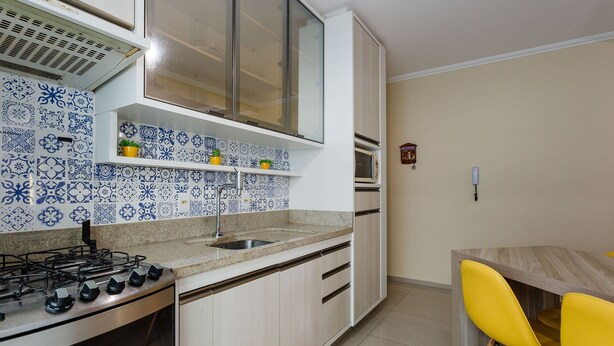 A kitchen with a gas stove, a sink, and a countertop. There is a cabinet with a glass door and a clock on the wall. - Beautiful Apt With BBQ 850m From the Beach Ral204 (Florianópolis)
