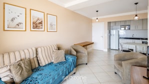 A living room with a sofa, armchair, and a small wooden table. There is a kitchen area visible in the background. - Apart w/ Pool 400m Praia dos Ingleses Posc0311 (Florianópolis)