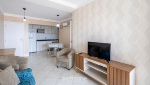 A modern living room with a TV, sofa, and armchair. A kitchen is visible in the background. - Apart w/ Pool 400m Praia dos Ingleses Posc0311 (Florianópolis)