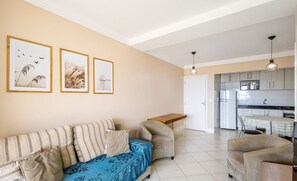 A living room with a sofa, armchair, and a small wooden table. There is a kitchen area visible in the background.