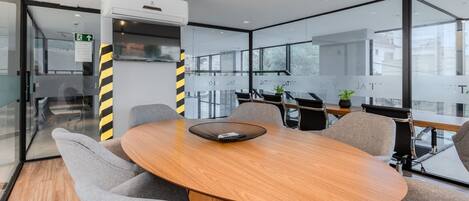 A modern meeting room with a round wooden table, grey upholstered chairs, and a glass partition with windows.