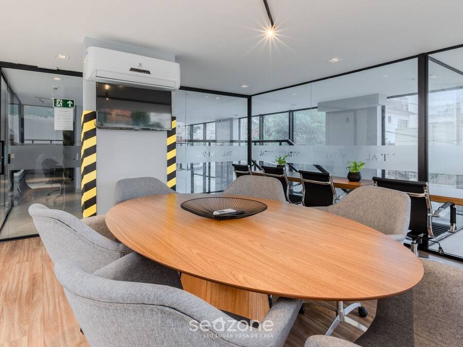 A modern meeting room with a round wooden table, grey upholstered chairs, and a glass partition with windows.